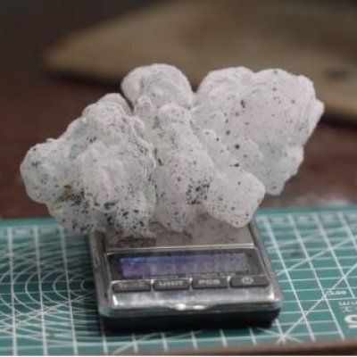 A spiky white hailstone, flecked with small dots of black paint, rests on a scale on a desk. 